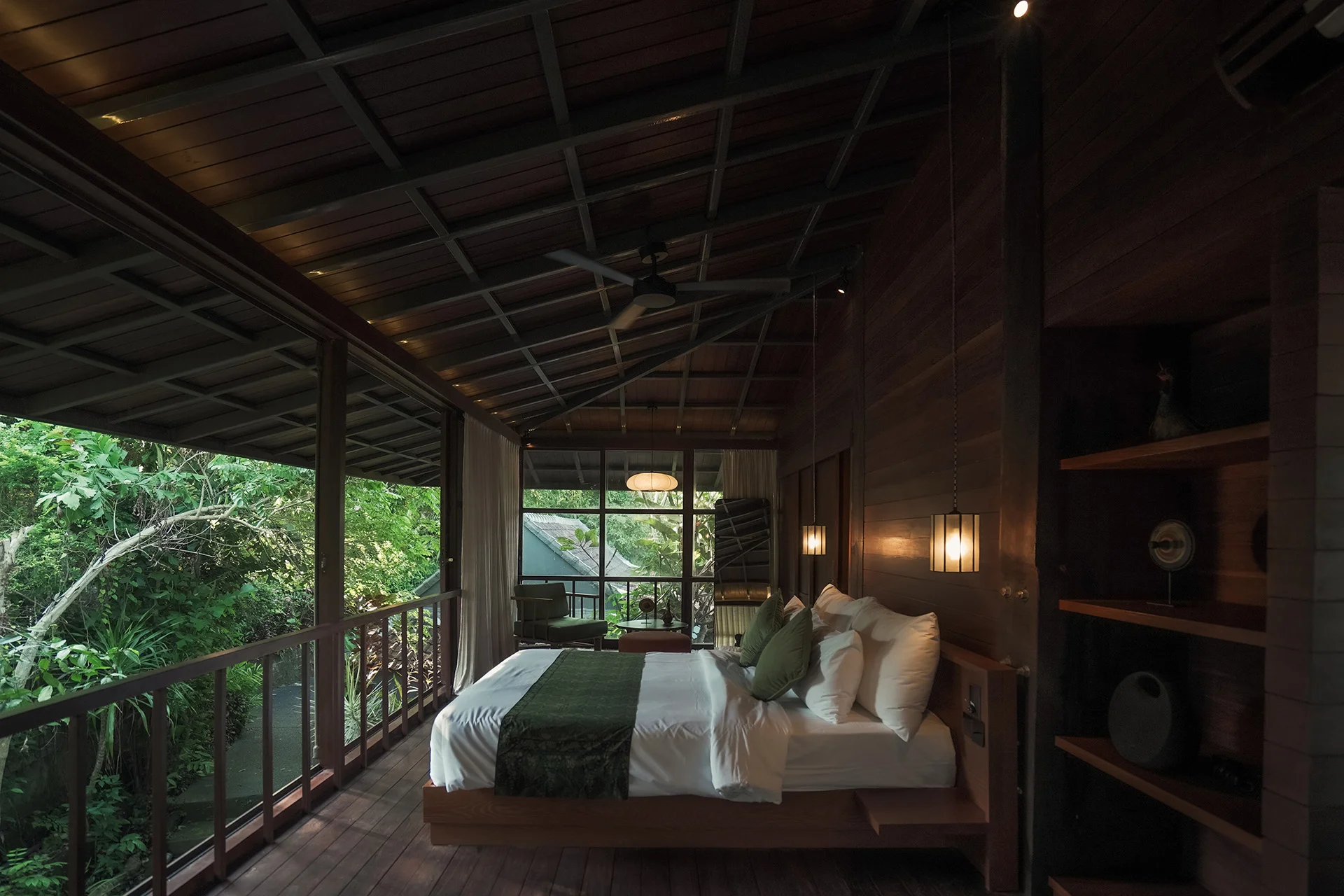 The treehouse bedroom, a low bed with green and white linen beside an open balcony rail, timber shelving on the wall