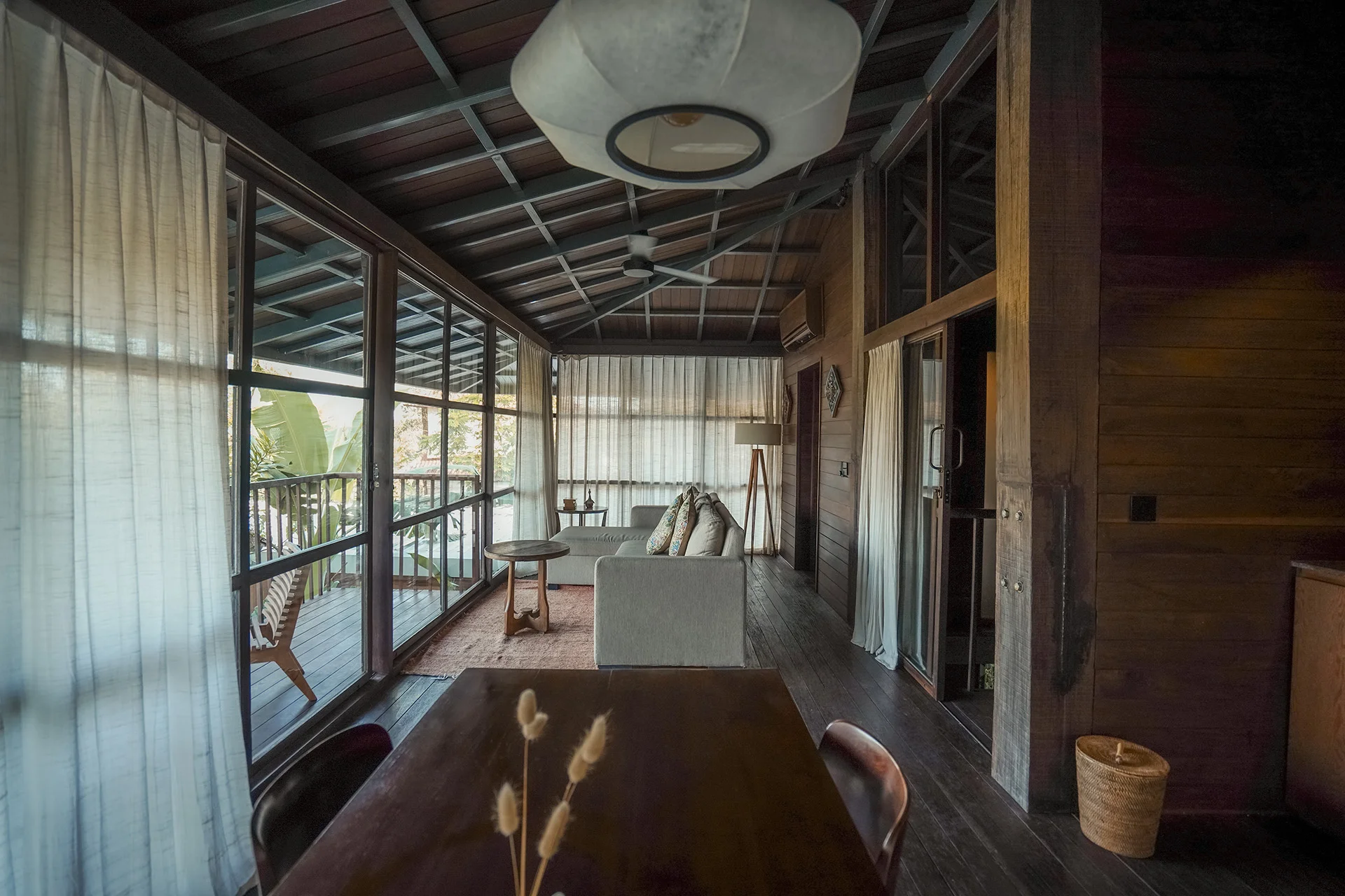 The treehouse dining table and sofa along a glass wall with white curtains, a pendant lamp above and the trees outside